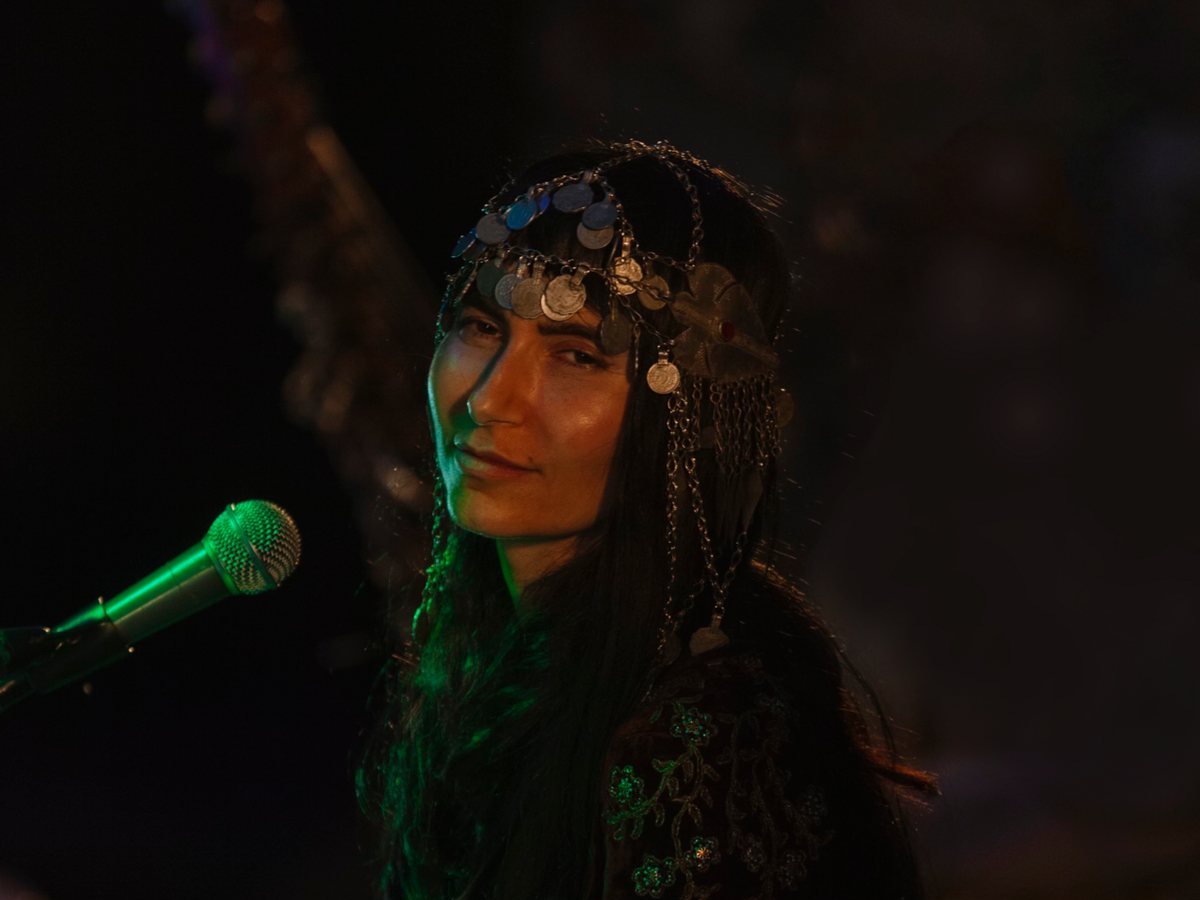 Simrit on stage beside a microphone in a coin headdress, 2021