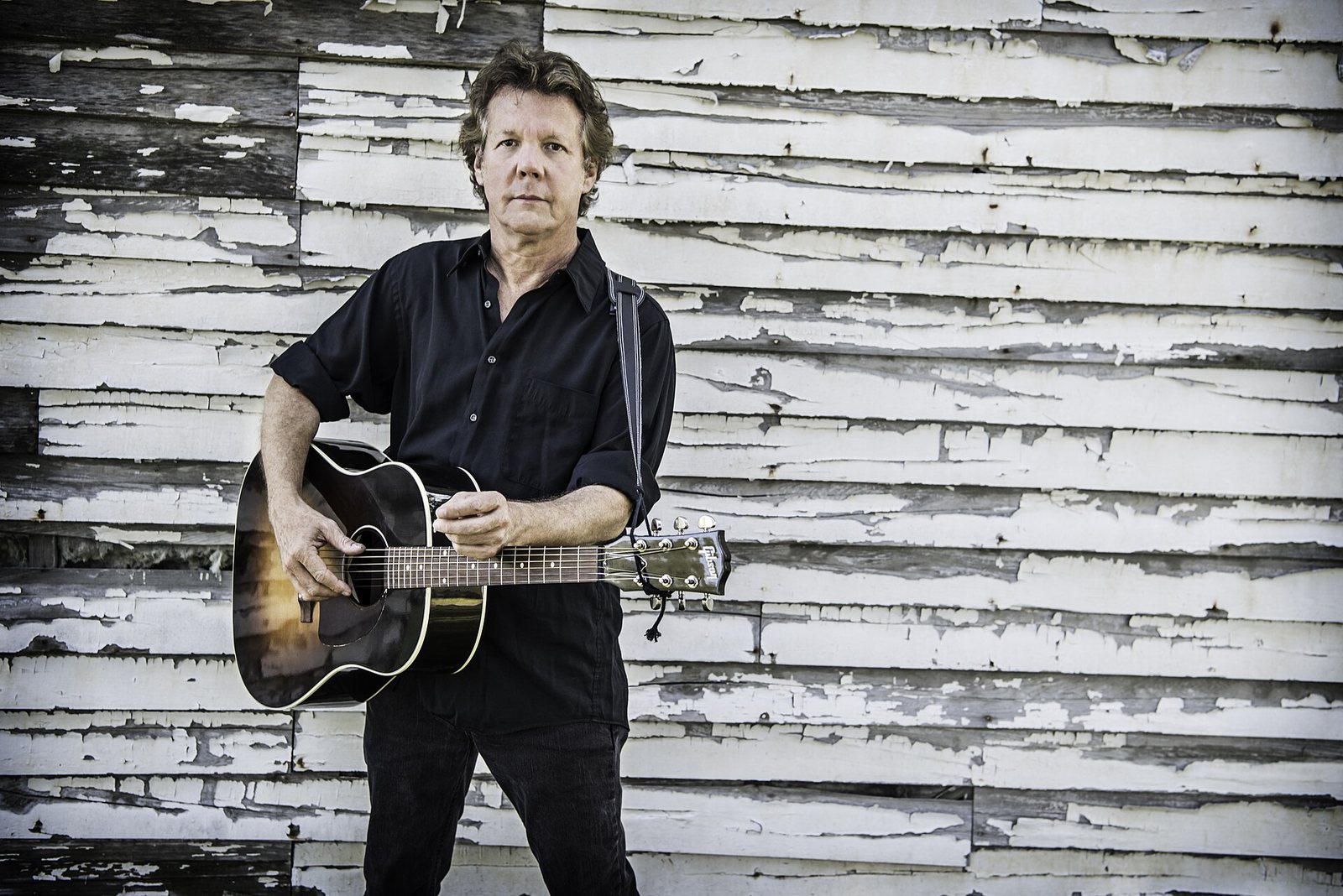 Steve Forbert with an acoustic guitar against a weathered white wooden wall, 2016