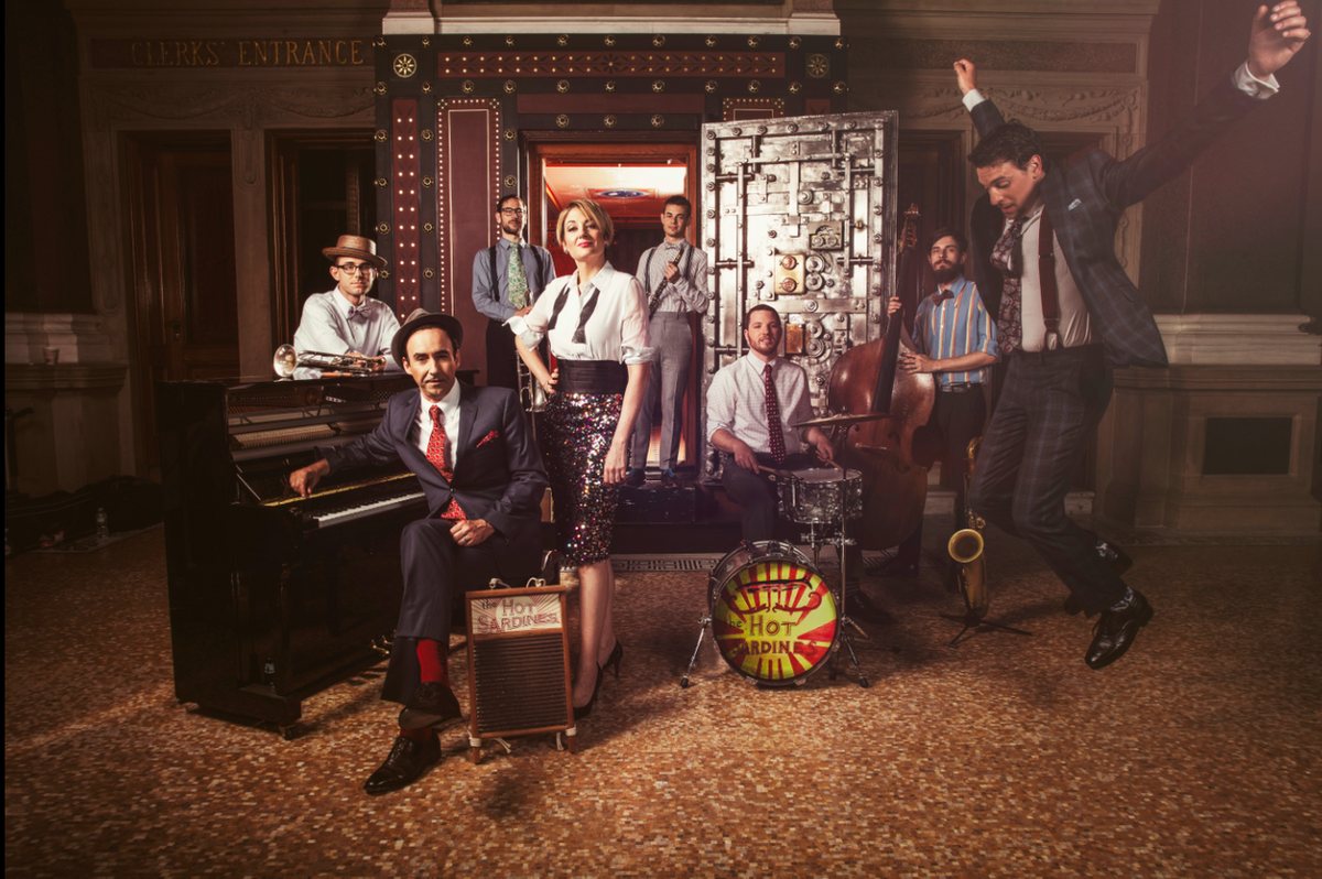 The Hot Sardines posing with piano, drums and upright bass beside a vintage bank vault door