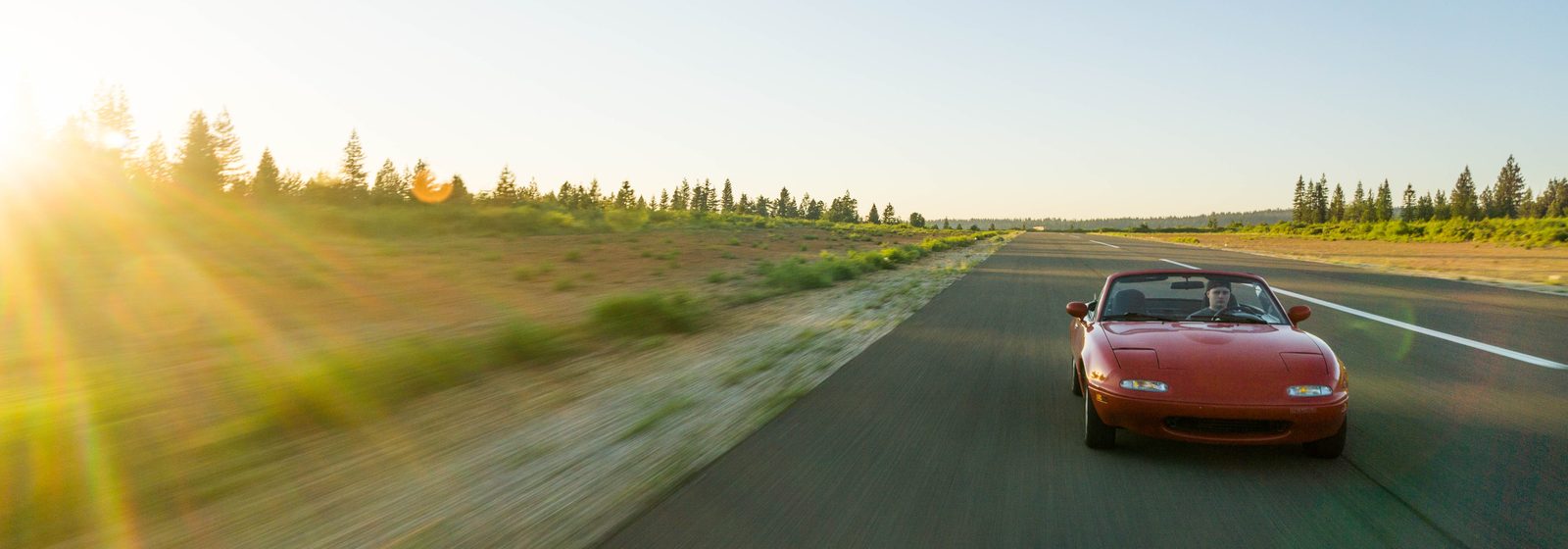 Red convertible driving on an open highway lined with pine trees at sunset