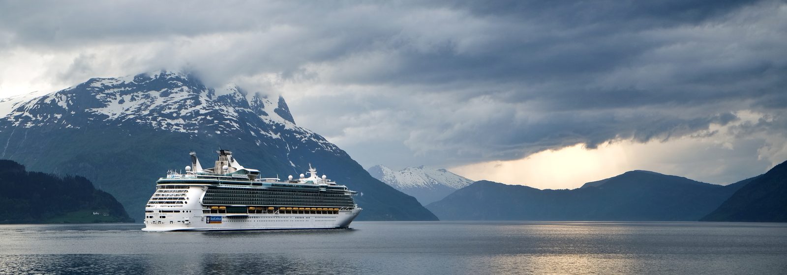 Cruise ship sailing through a Norwegian fjord beneath snowy peaks and storm clouds