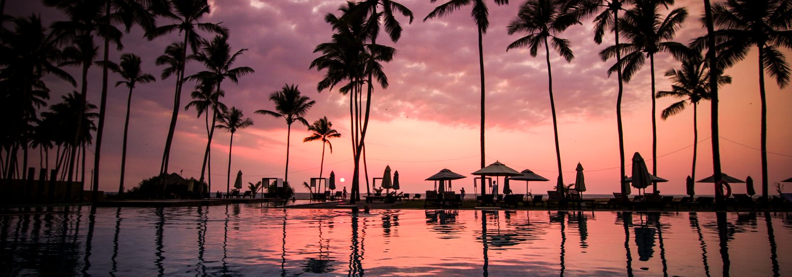 Silhouetted palm trees and beach umbrellas reflected in a resort pool at sunset