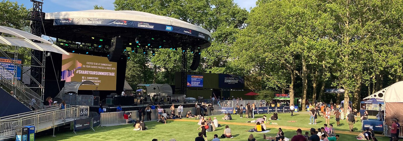 Wide view of the Central Park SummerStage outdoor stage and lawn in Manhattan, New York City, with concertgoers seated on the grass - CityParks Summerstage tickets
