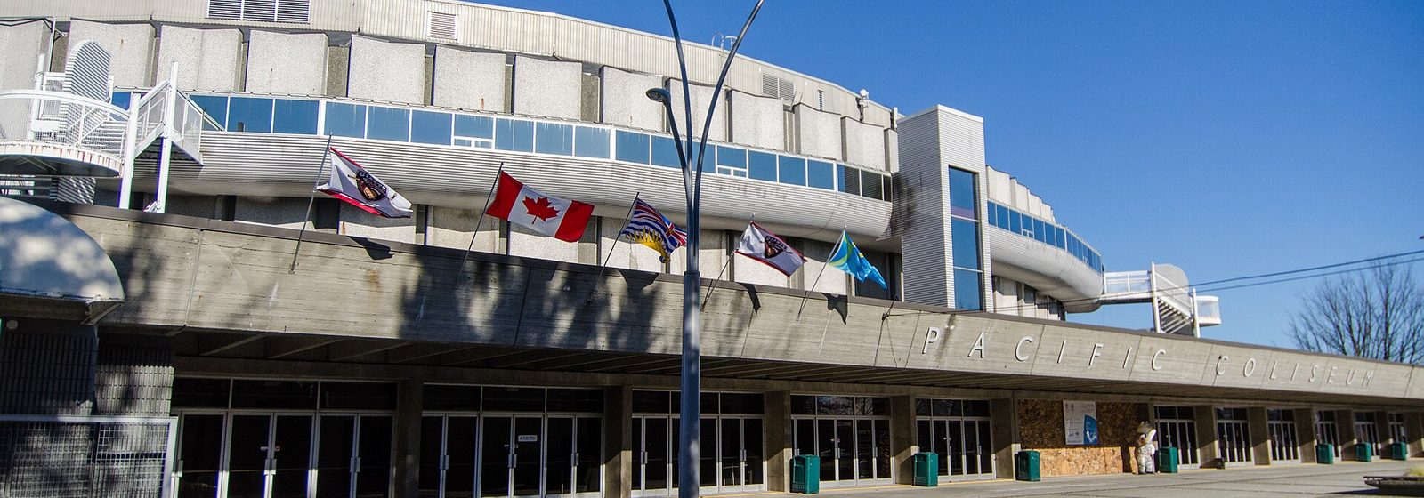 Exterior facade of the Pacific Coliseum in Vancouver, British Columbia, with flags flying above the entrance plaza and the venue name lettered on the concourse - Pacific Coliseum tickets