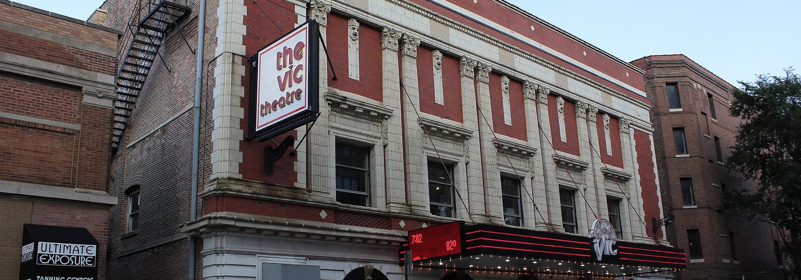 Street view of the brick and terra-cotta facade of the Vic Theatre in Chicago, with its vertical sign and lighted marquee above the box office entrance - Vic Theatre tickets